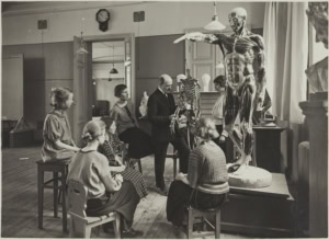 1920-luvun koululuokka, jossa joukko nuoria naisia ja miesopettaja tutkivat ihmisen luurankoa.